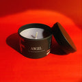 Candle in a black container labeled 'Angel' on a red background. BTS 
