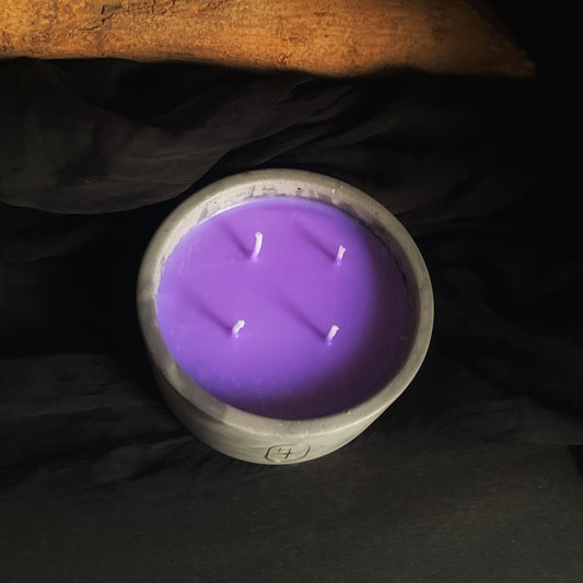 A purple soy wax candle is displayed in a concrete jar with a wooden background. The candle has multiple wicks and has a woody scent.