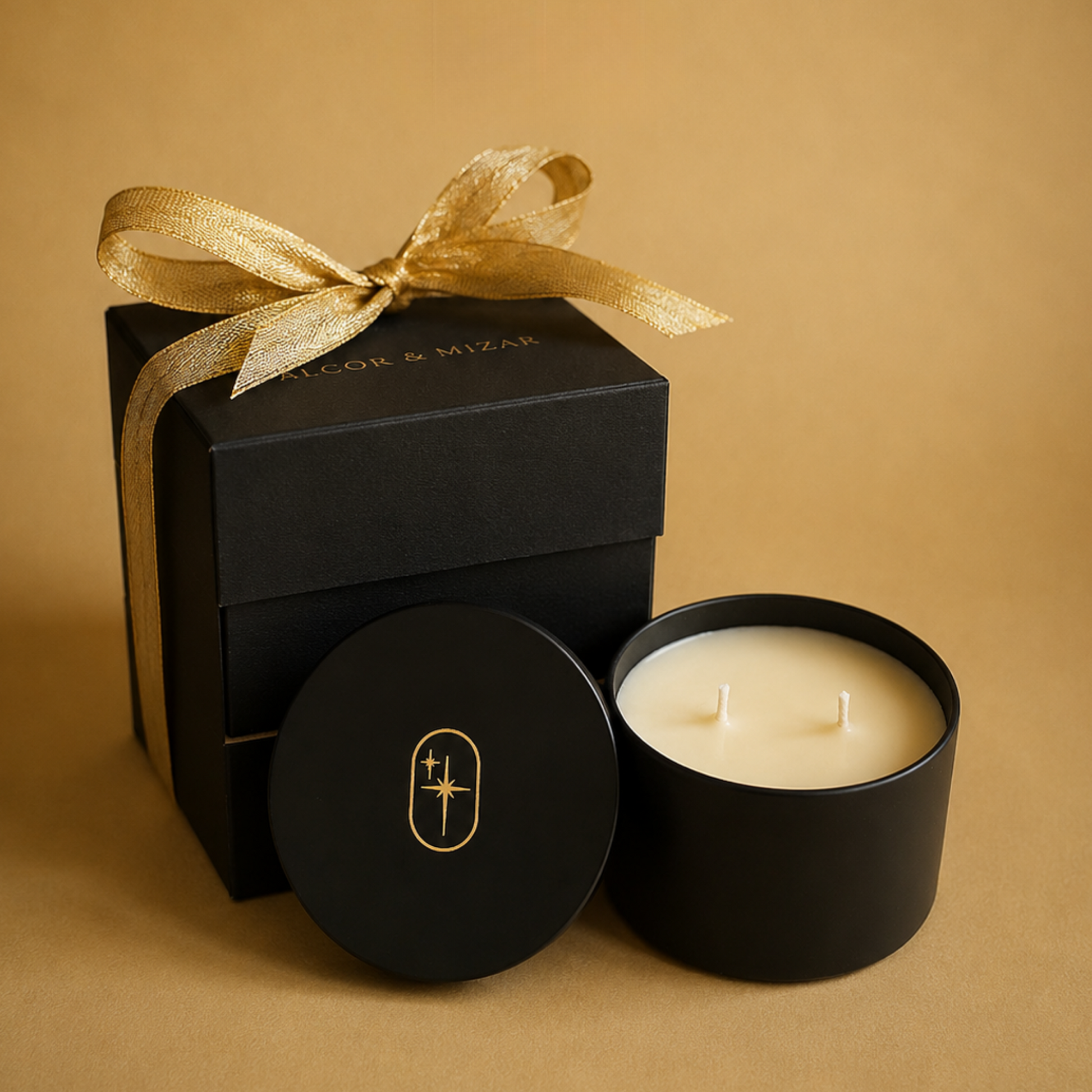 A black candle tin with cream-colored wax sits beside its matching black lid and a black gift box tied with a gold ribbon. The lid features a small gold logo, and the box has subtle gold text, all set against a warm tan background.
